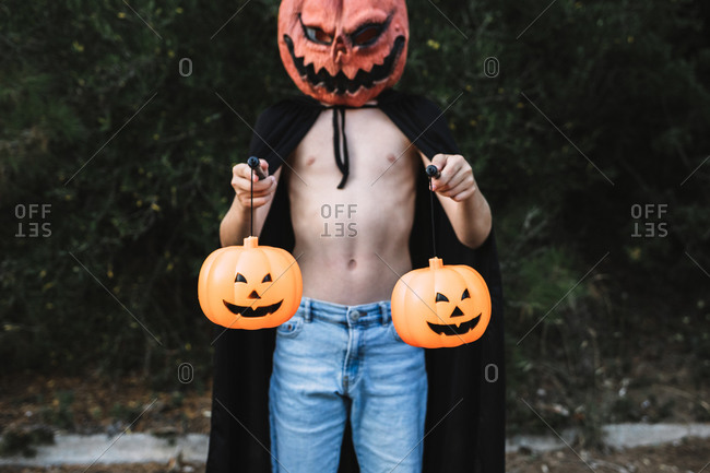Unrecognizable male wearing creepy mask and black cloak standing with pumpkin lanterns on city street