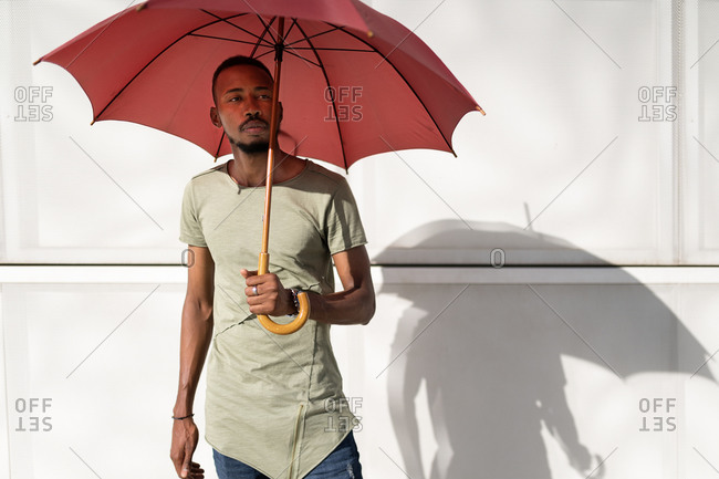 Tranquil black male standing under umbrella in city looking away and enjoying sunny weather in summer