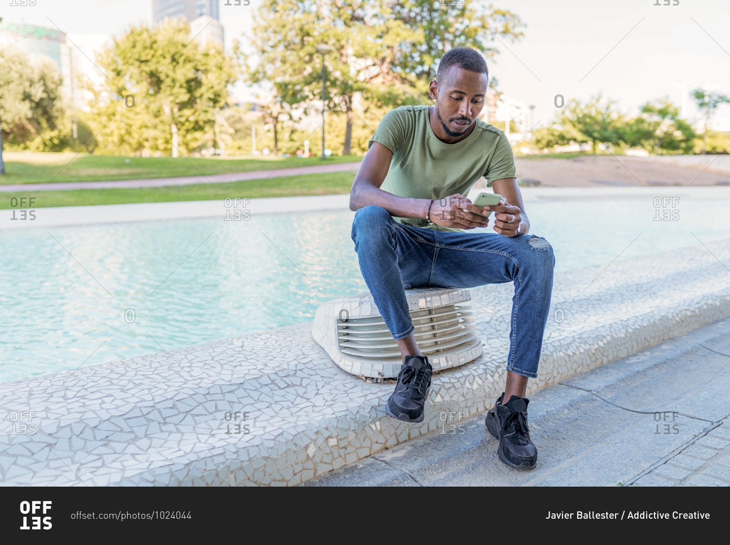 Pensive African American male sitting on border near fountain in city and surfing Internet on smartphone while chilling during