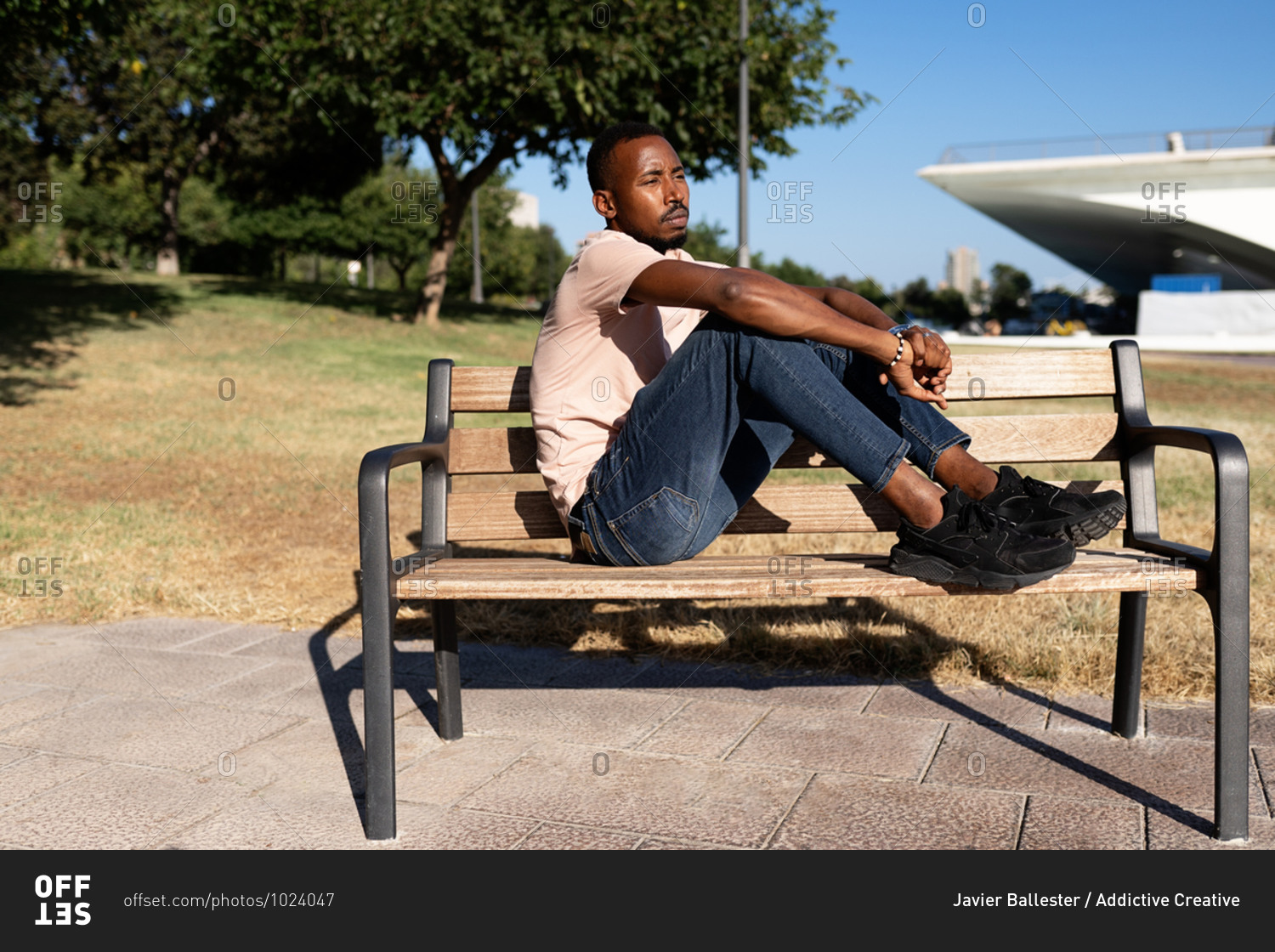 Side view of relaxed African American male sitting on bench in urban park and embracing knees while looking away