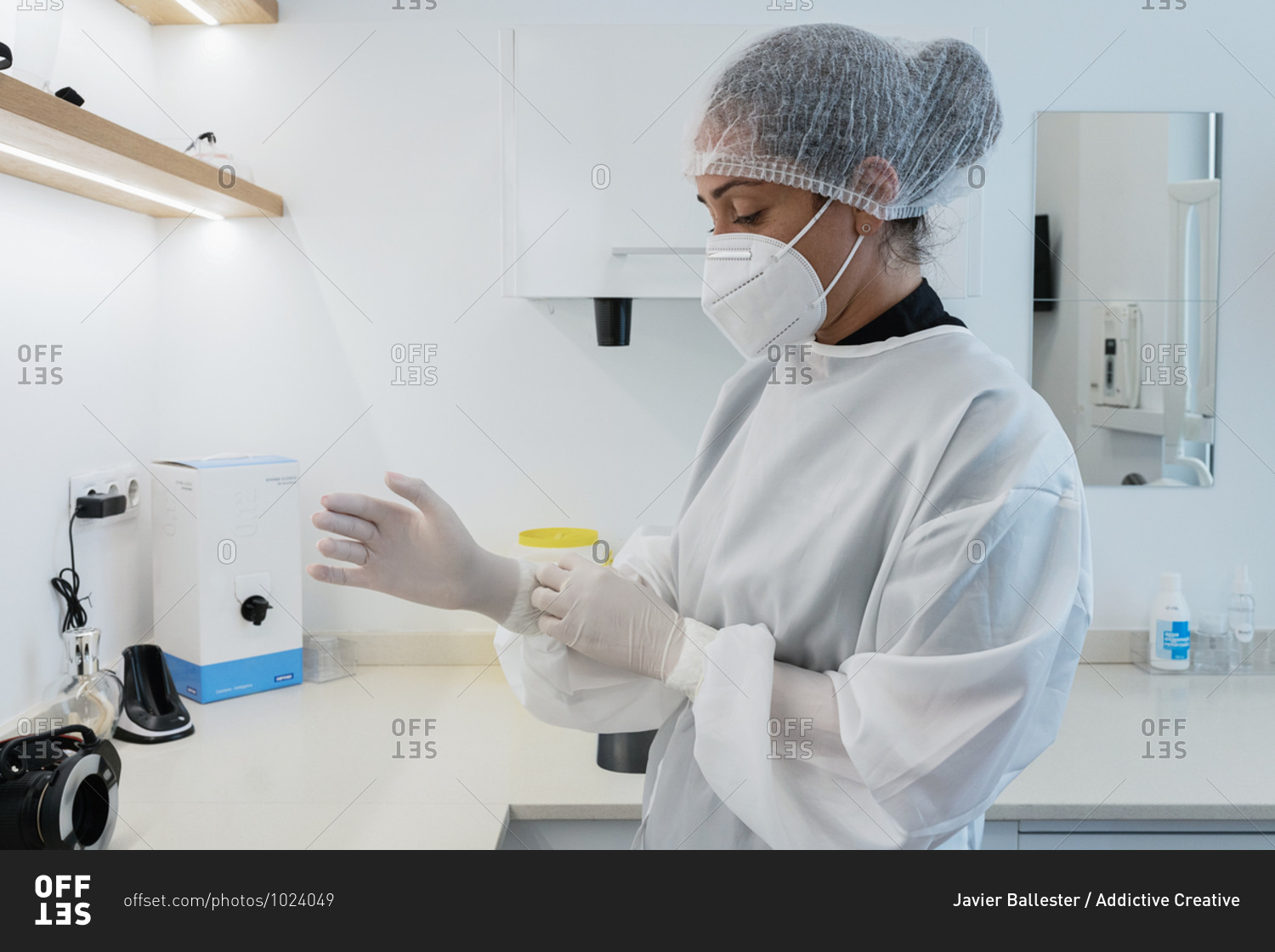 Side view of young female medical worker in white uniform and mask putting on latex gloves while preparing for treatment in modern clinic