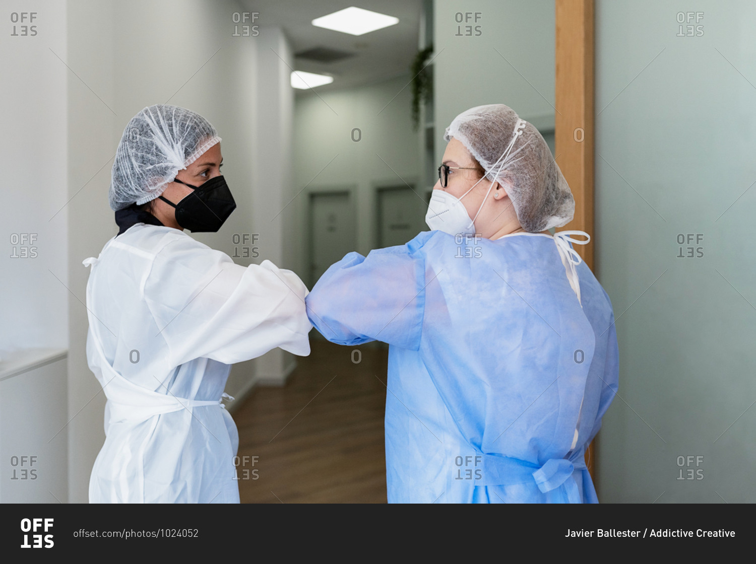 Back view of female medical workers in uniform and protective masks greeting each other with elbow bump in clinic during coronavirus pandemic