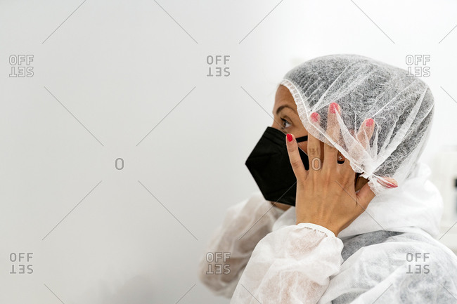 Side view of female medical specialist putting on protective suit with face mask and protective cap while preparing for work during coronavirus pandemic