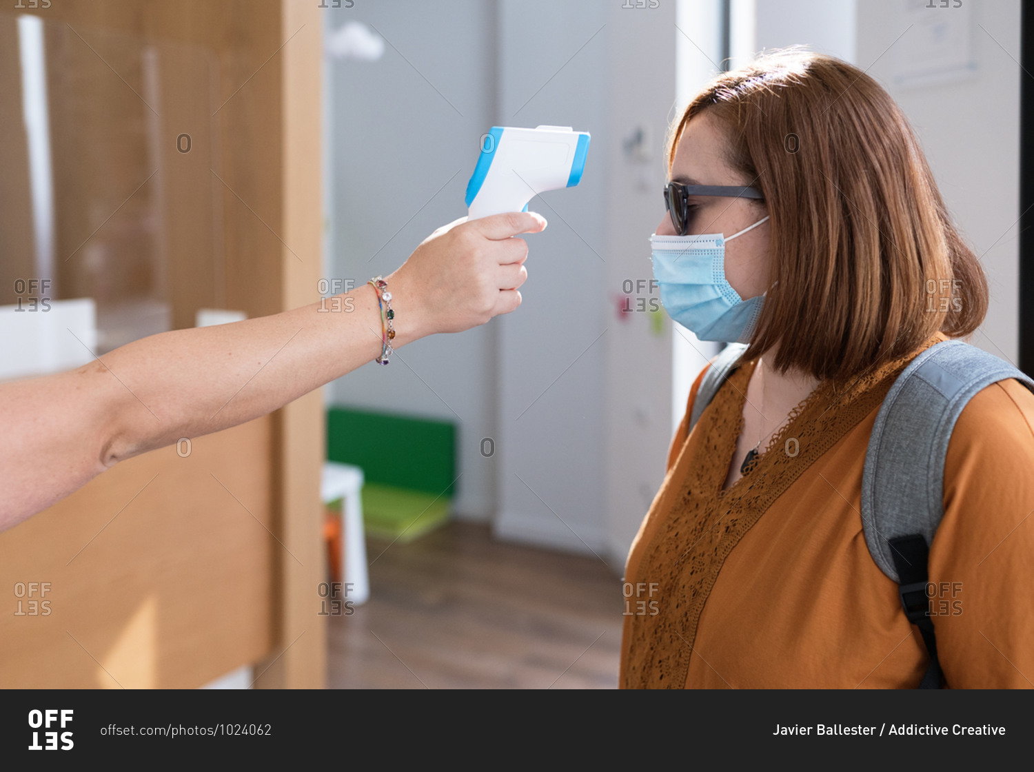 Side view of anonymous specialist with digital thermometer gun measuring temperature of female visitor in protective mask before entering clinic