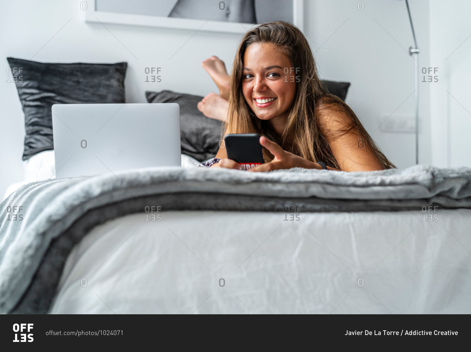 Positive female in nightwear lying looking at camera on soft bed and reading messages on smartphone in morning after sleep