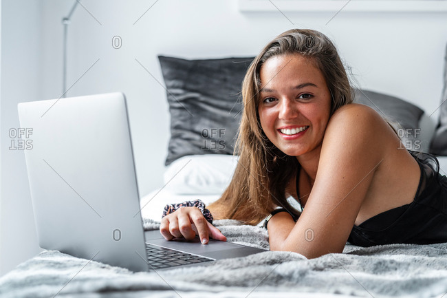 Delighted female in sleepwear lying looking at camera on cozy bed at home and typing on netbook in morning while chatting on social media