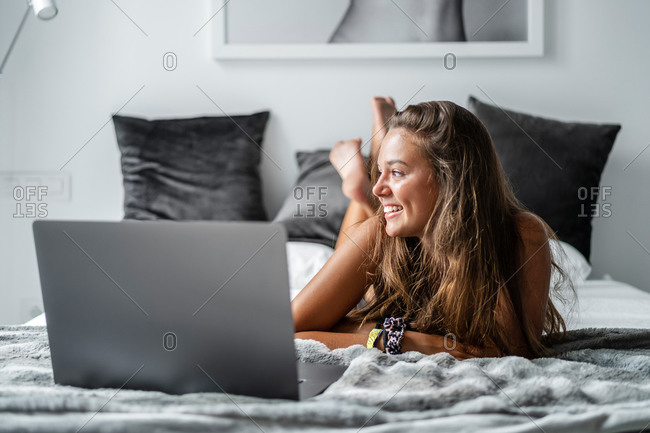 Delighted female in sleepwear lying on cozy bed at home and typing on netbook in morning while chatting on social media