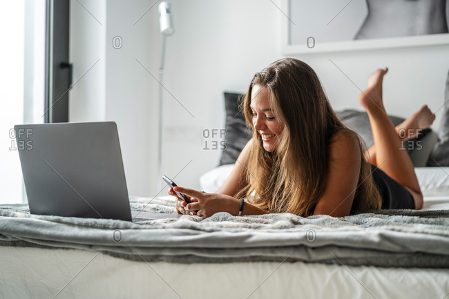 Positive female in nightwear lying on soft bed and reading messages on smartphone in morning after sleep