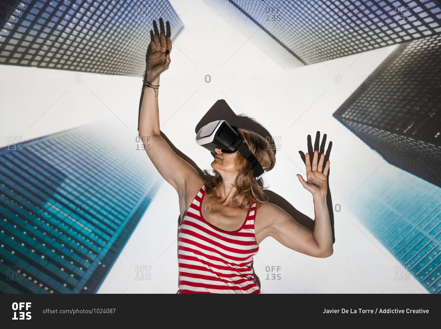 Unrecognizable female in casual wear and VR headset standing with arms raised against contemporary skyscrapers inside virtual city