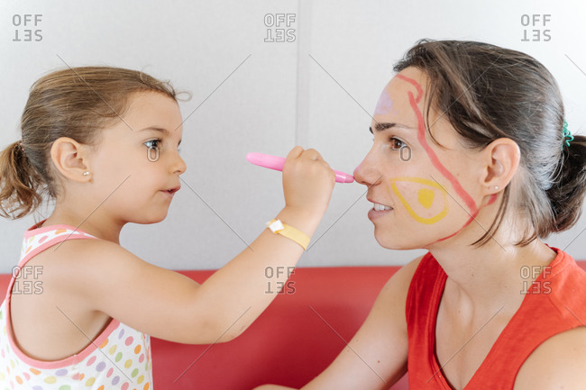 Side view of adorable little kid painting pictures on face of mother while entertaining during weekend at home