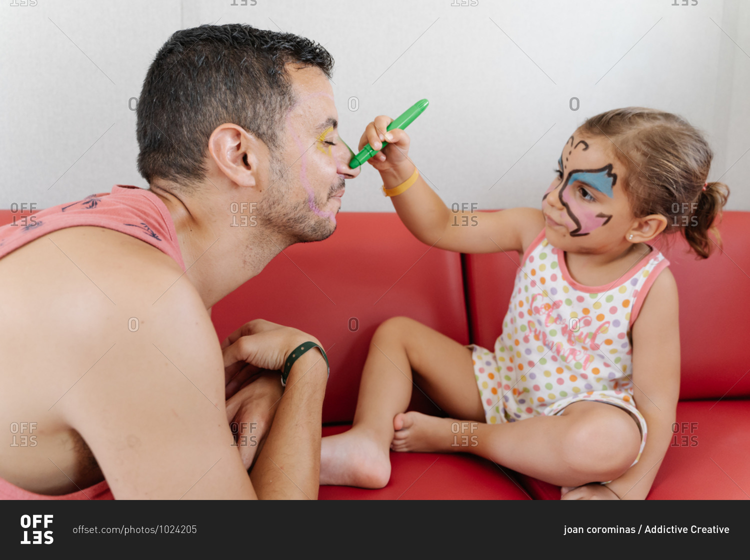 Side view of adorable girl painting face of calm father while relaxing together on sofa and having fun during weekend