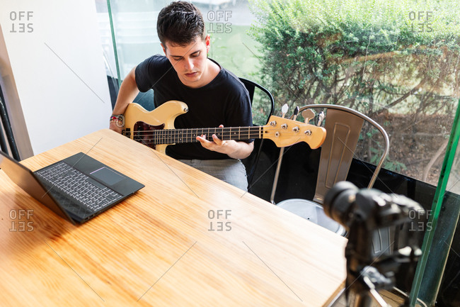 Focused young male musician sitting near window and performing music on bass guitar