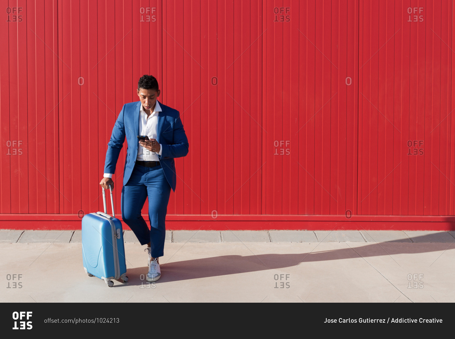 Full body of young African American man in elegant blue suit with luggage messaging on mobile phone while standing against red wall on street