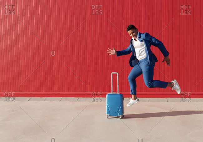Full length side view of happy young African American male entrepreneur in stylish outfit with suitcase screaming and jumping near red wall while celebrating business success