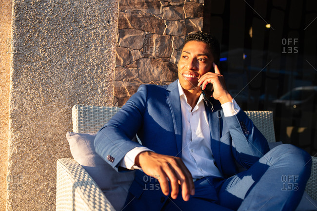 Young successful well dressed black businessman in elegant suit sitting in chair and having pleasant conversation on mobile phone