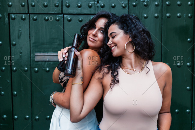 Positive relaxed young Latin girlfriends clinking bottles of beer while chilling together against green wall