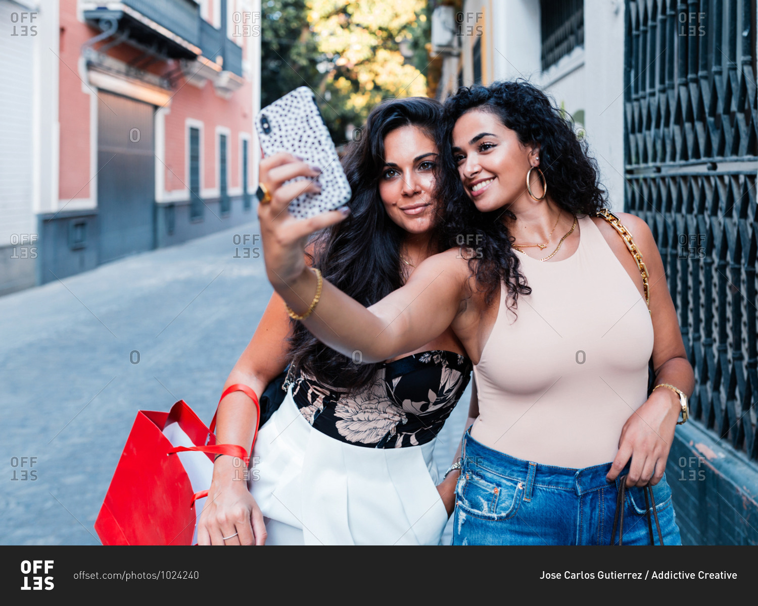 Cheerful young Latin female friends in stylish clothes standing on narrow street and taking selfie with smartphone while spending summer day together in city