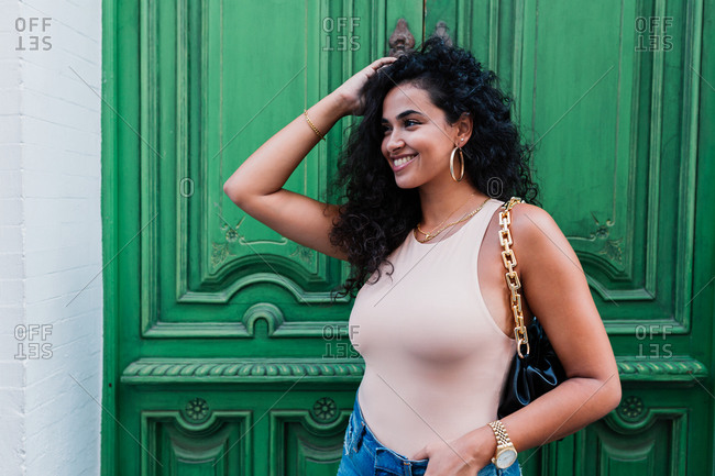Cheerful young Latin female with long curly hair in casual outfit and with accessories looking away and smiling happily while standing against weathered green door of old building