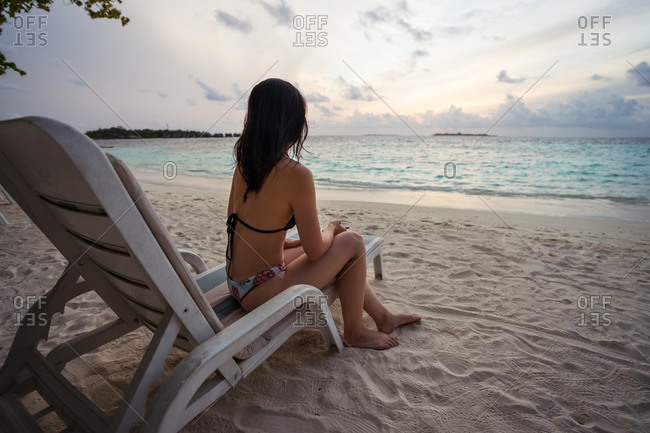 Back view of unrecognizable female tourist in bikini sitting on chair and enjoying amazing sunset over waving sea while resting on sandy beach during summer vacation on Maldives islands