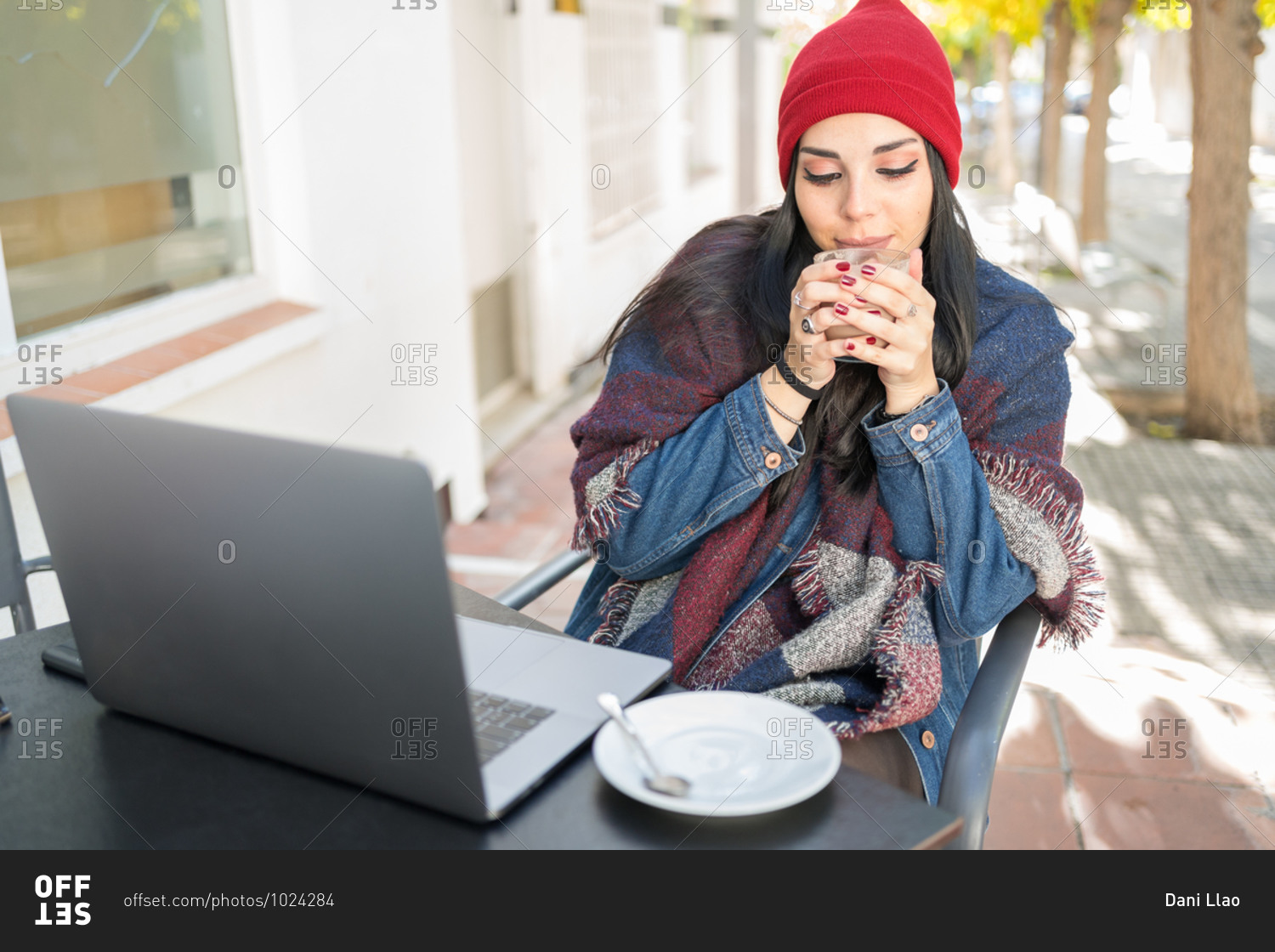 Young brunette woman warms her hands with a hot cup of coffee on the terrace of a bar protected by a blanket while she checks her laptop