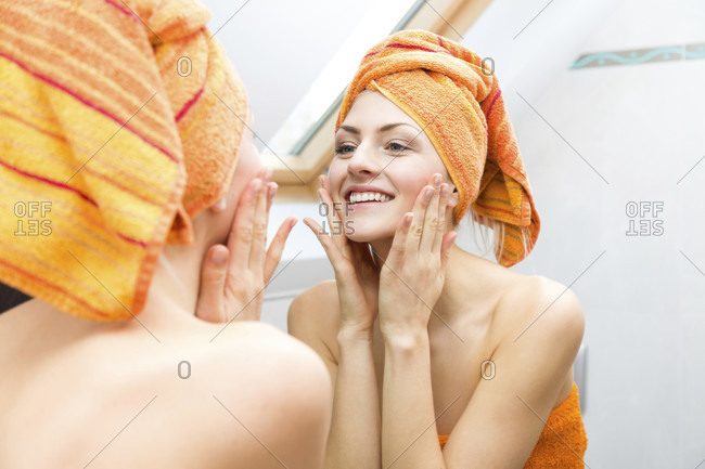 Attractive woman wrapped in towel touching the sides of her face with big happy smile in reflection on mirror