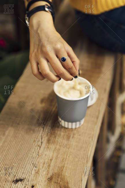 Hand of woman stirring coffee in disposable cup on wooden bench