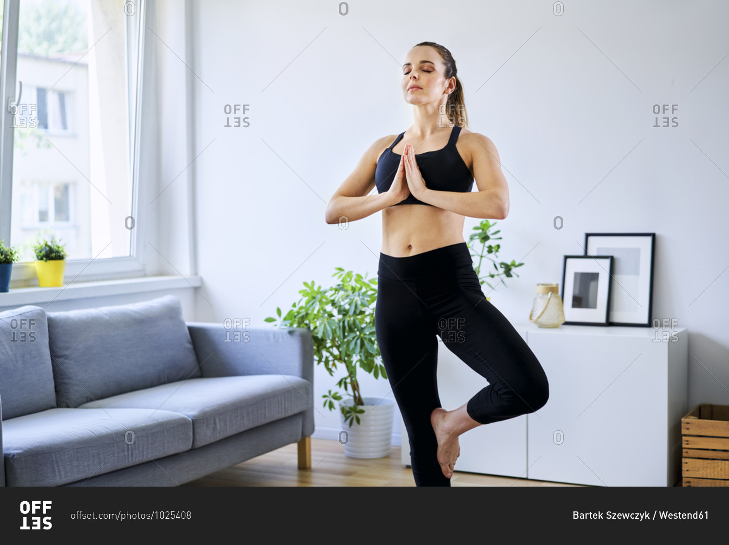 Woman practicing tree pose with eyes closed in living room Stock