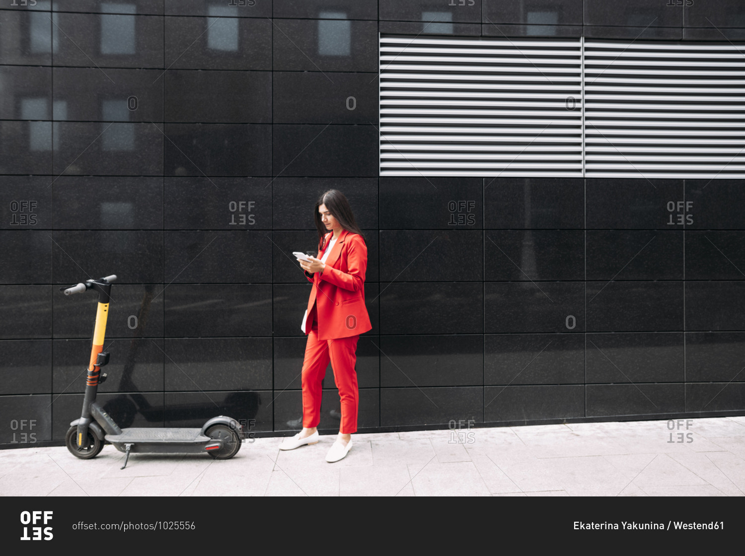 Female entrepreneur in red business wear using smart phone while standing against black building in city