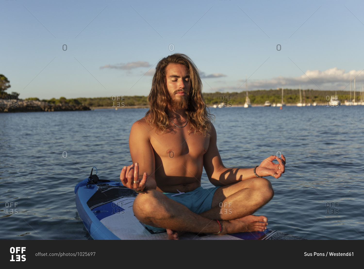 Young man with long hair meditating while sitting on paddleboard at sea against sky during sunset