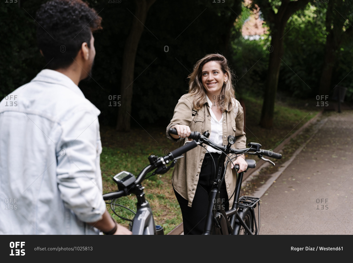 Smiling young woman looking at boyfriend while standing with electric bicycle on road