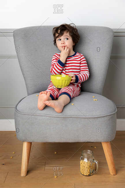Young boy sitting on chair eating cereal with bowl on his lap