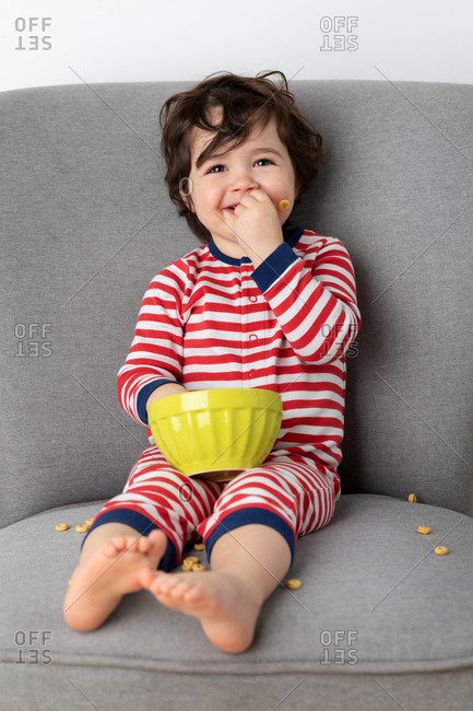 Smiling toddler sitting on sofa eating cereal with hands