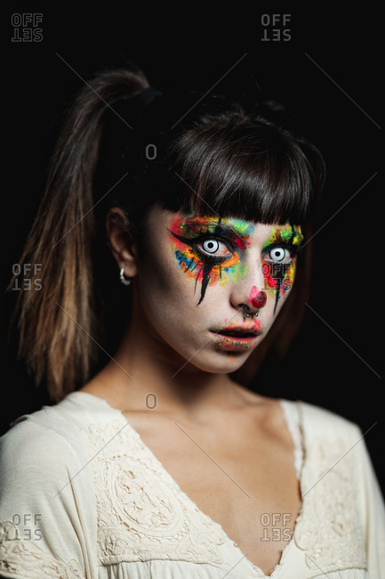 Scary young female in colorful contact lenses and with painted face standing in dark forest and smiling creepy while looking at camera on Halloween