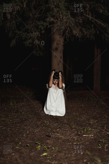 Spooky female sitting on ground in woods at night and touching hair while looking at camera on Halloween