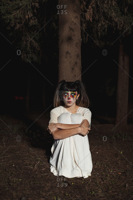 Spooky female sitting on ground in woods at night looking at camera on Halloween