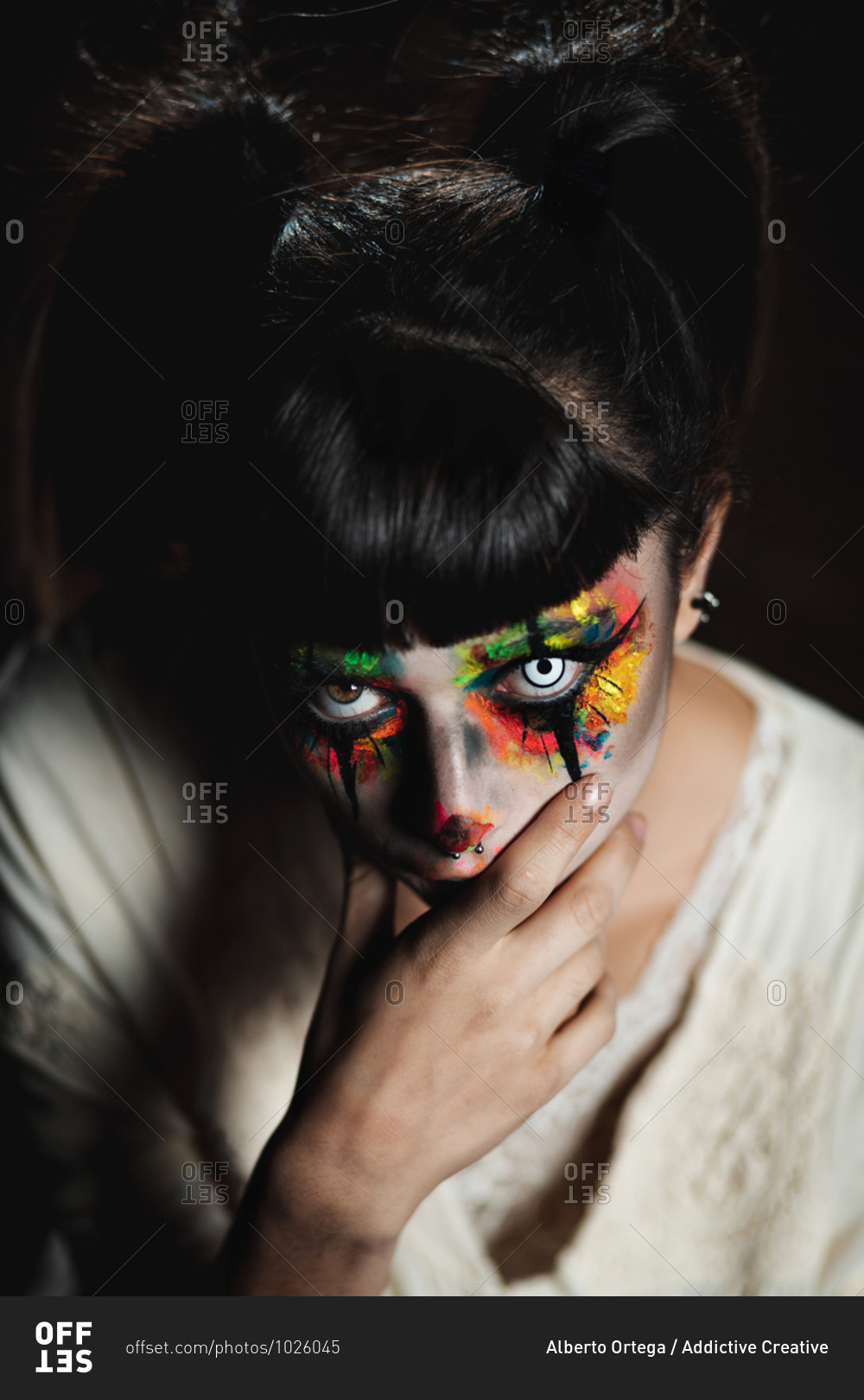 Scary young female in colorful contact lenses and with painted face  standing in dark forest and