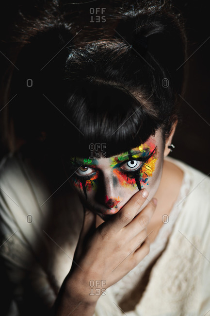 Scary young female in colorful contact lenses and with painted face standing in dark forest and smiling creepy while looking at camera on Halloween