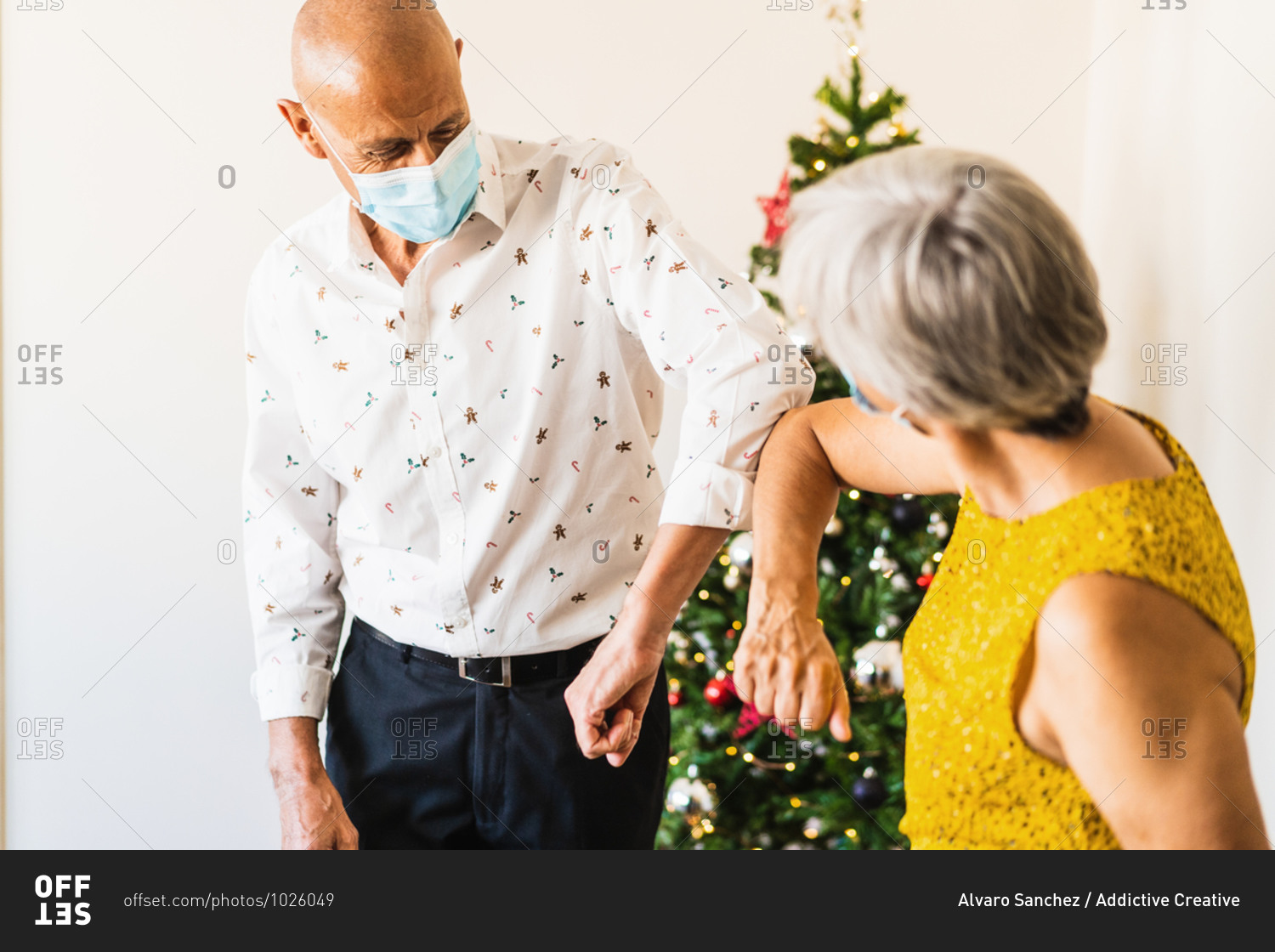 Positive mature couple in smart casual outfits and face masks greeting by bumping elbows while standing near decorated shiny Christmas tree