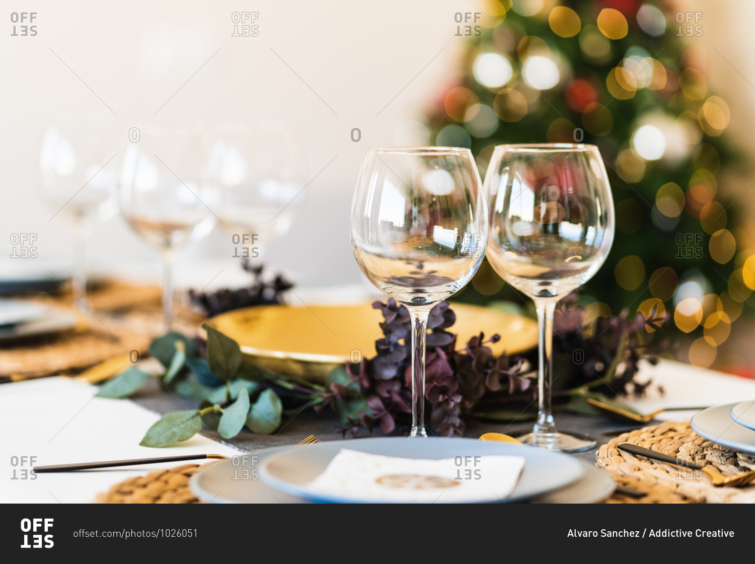 Dining table served with glasses and plates against blurred glowing Christmas tree on Christmas eve at home