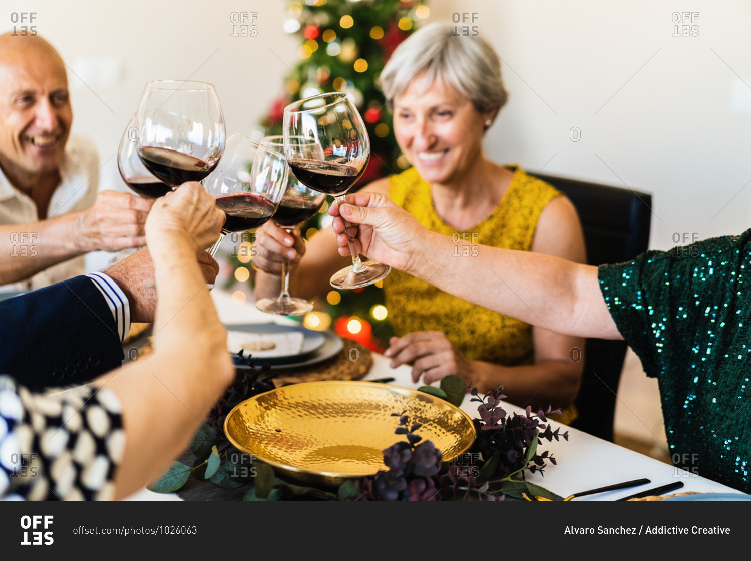 Happy mature friends gathering at festive table near shining Christmas tree and clinking wineglasses while celebrating Christmas eve together