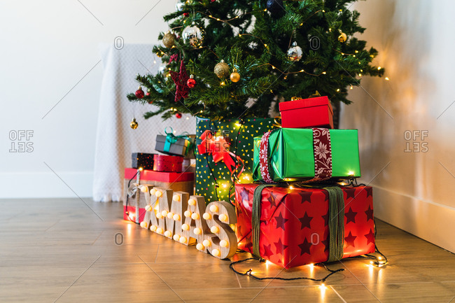 Heap of colorful ornamental present boxes with ribbons near decorative fur tree and shiny letters on floor with garland during festive event