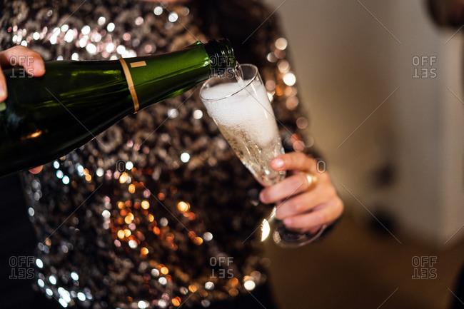 Unrecognizable female in elegant shiny dress standing in room and pouring cold champagne in glass during Christmas party