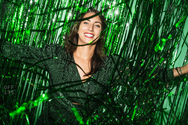 Delighted female wearing festive dress playing with green shiny tinsel stripes at party while looking at camera