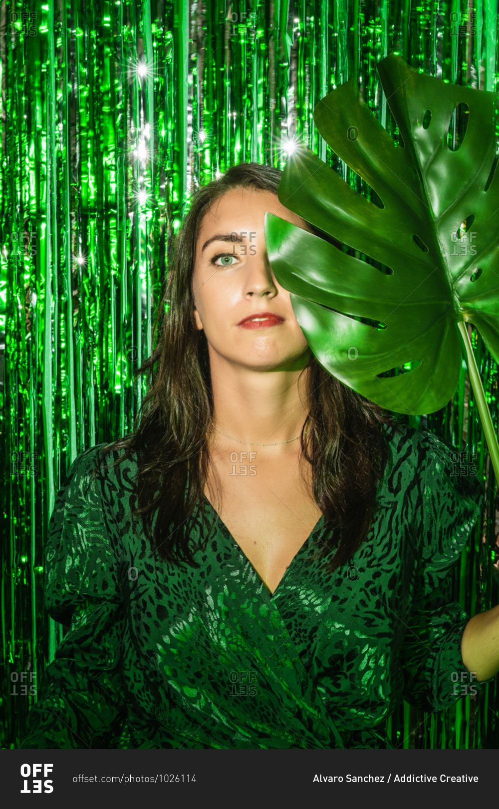 Calm female wearing stylish green dress standing with monstera leaf on background of shining foil tinsel stripes and looking at camera