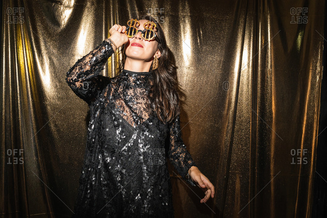 Content female wearing black glamour dress and fancy sunglasses having fun at party while standing on golden background
