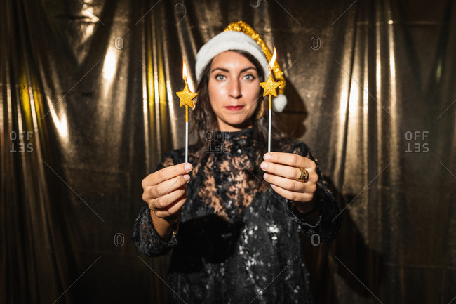 Calm female in Santa hat and fancy dress standing with burning candles and looking at camera while enjoying New Year party