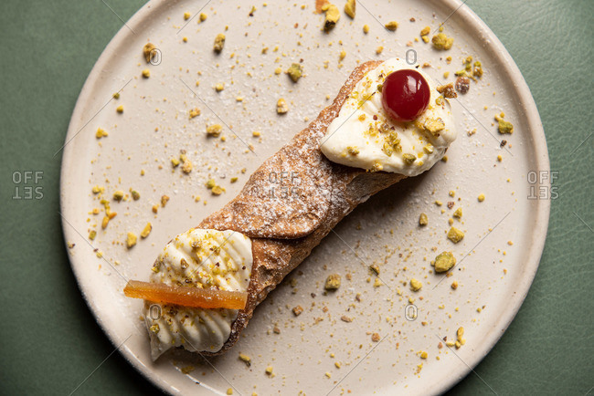 Overhead view of a cannoli on a plate garnished with cherry and pistachios
