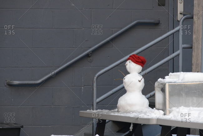 Small snowman with a red hat on a table outside gray building