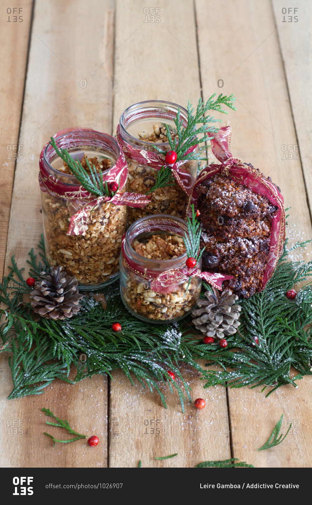 Glass jars filled with granola and decorated with green herbs and red berries arranged on wooden table with pine cones for Christmas celebration