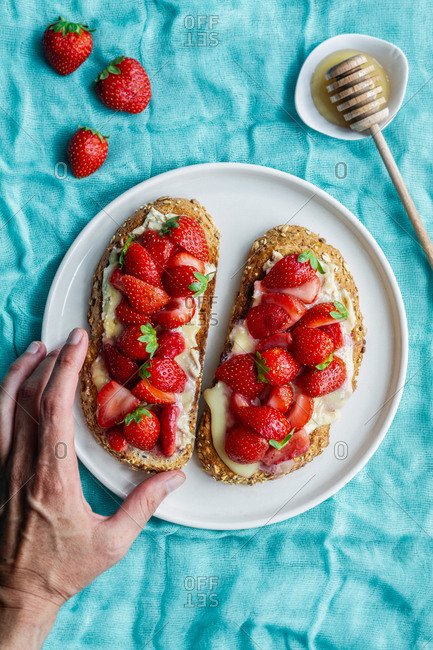 Top view of unrecognizable woman taking a yummy sweet bread toasts with fresh chopped strawberries and honey served on plate on blue tablecloth